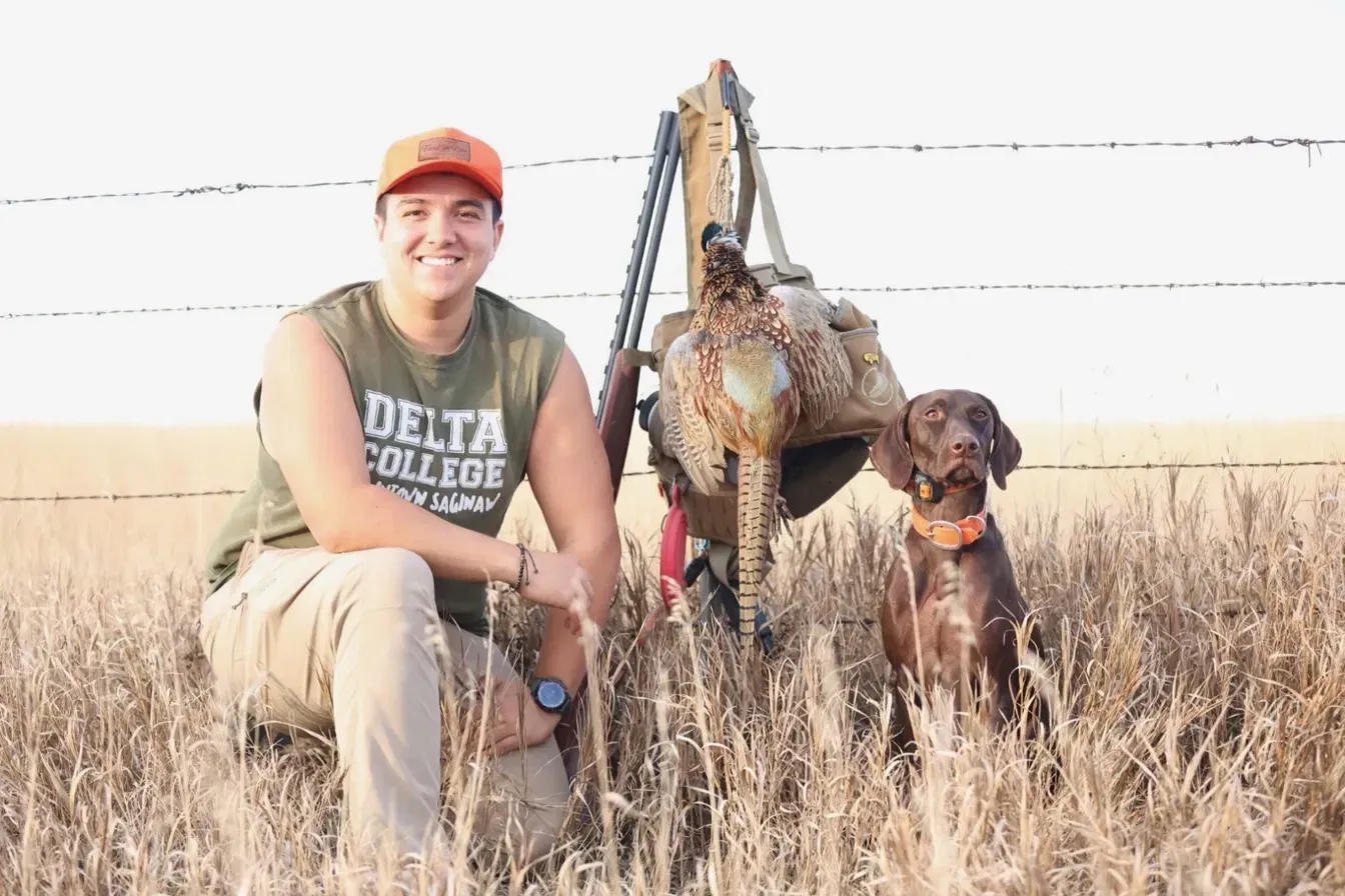A man kneeling in tall dry grass beside a brown dog sitting at attention.