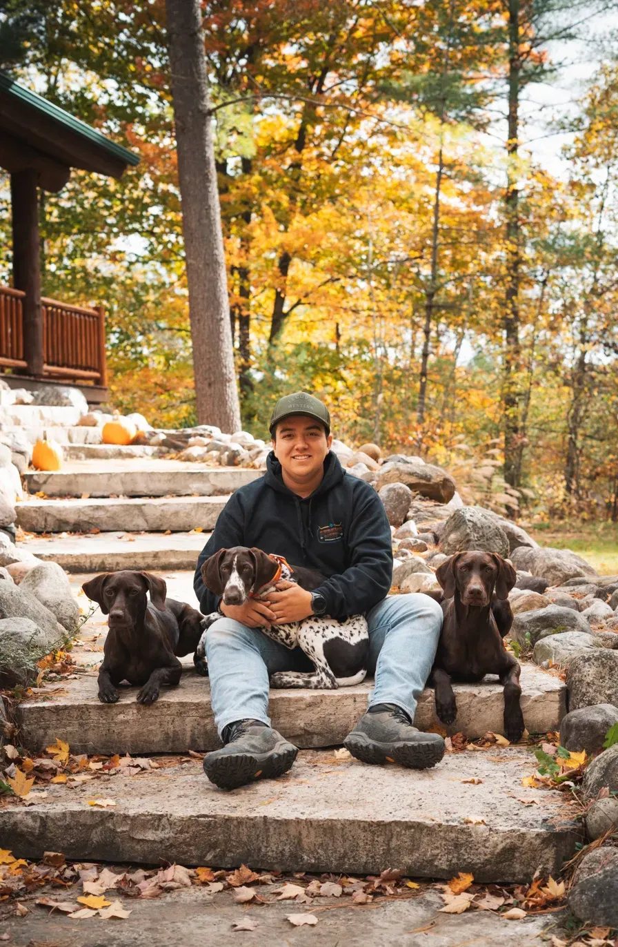 A man sitting on stone steps in autumn woods with three dogs.